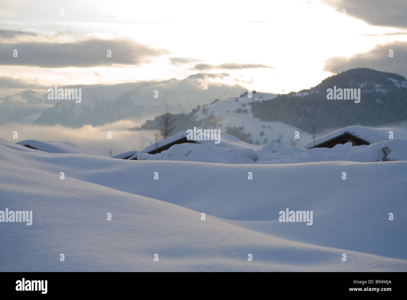 Un banco de nieve o se desvía en primer plano los techos cubiertos de nieve y montañas de fondo con la puesta de sol y nubes en el cielo Foto de stock