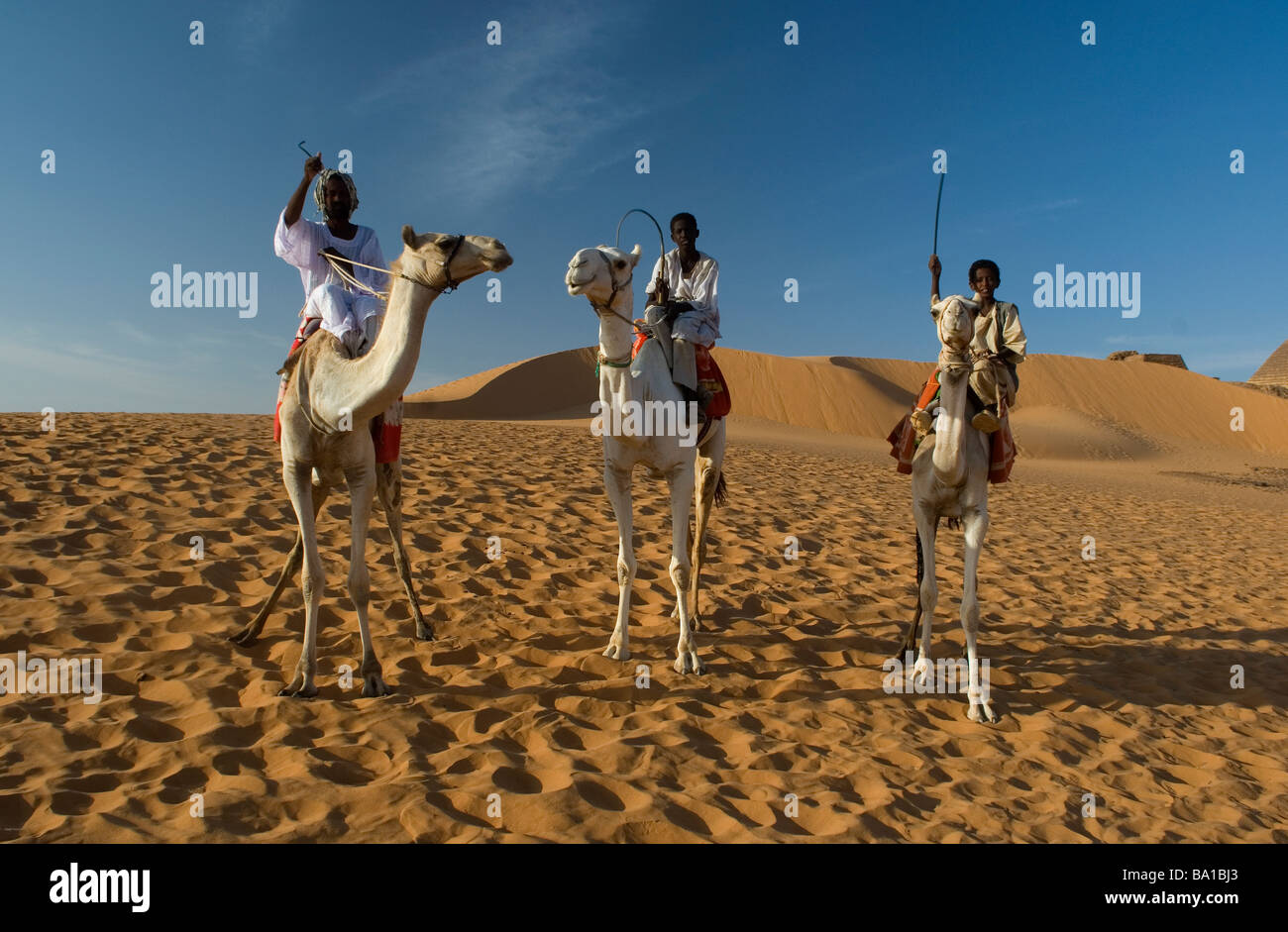 Los jinetes de camellos cruzan las dunas cerca de Merowe pirámides Foto de stock