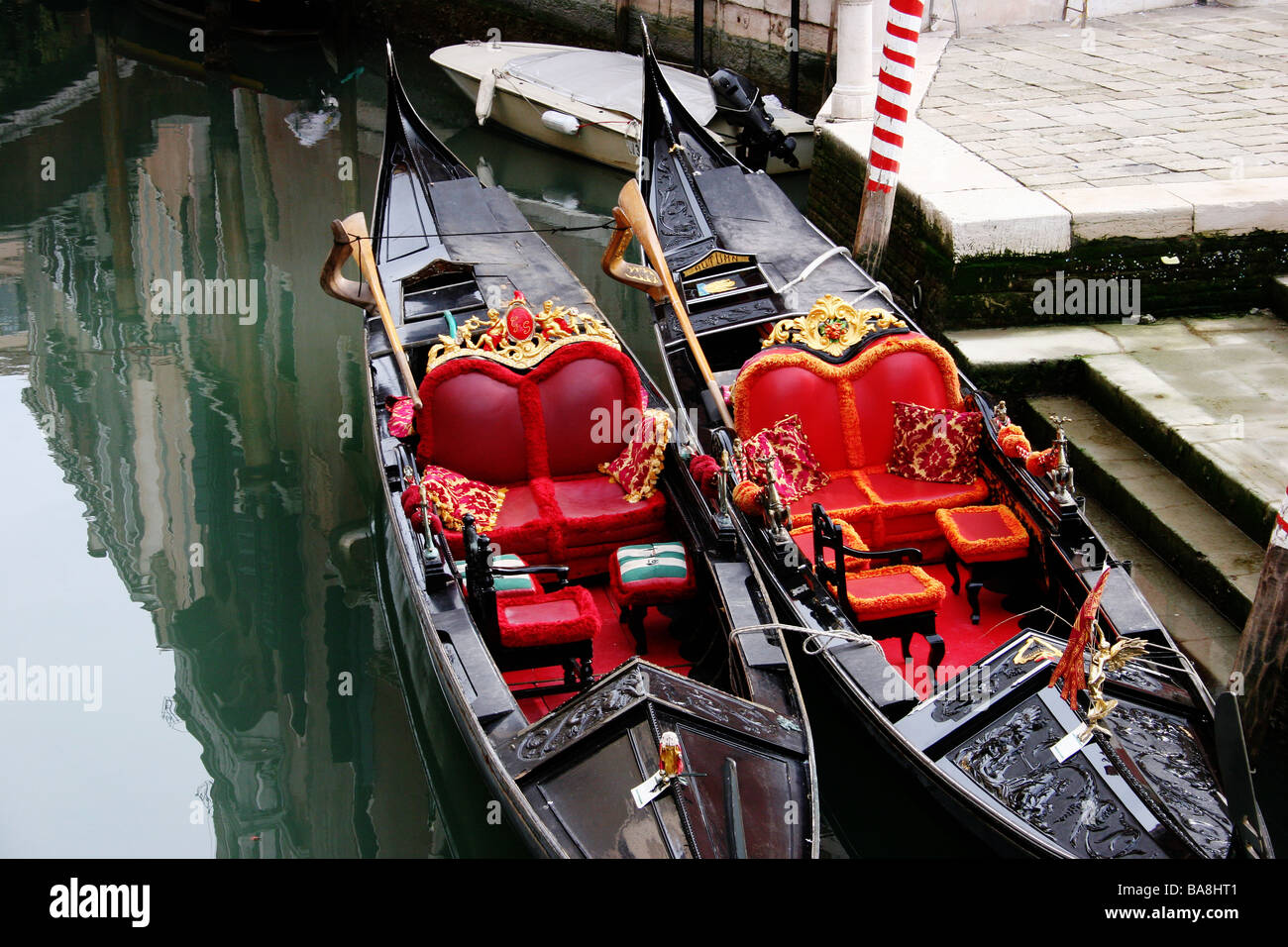 Dos Góndolas con asientos decorativos de color rojo, al lado de un canal con reflejan edificios venecianos Venecia Italia Foto de stock