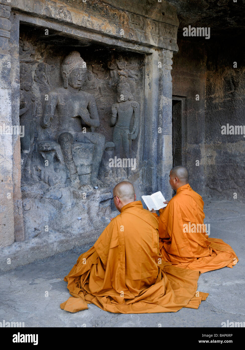 Monjes budistas en túnicas naranja sentados delante de la roca cortada Frescoe Buda orando en las Cuevas de Ajanta Foto de stock