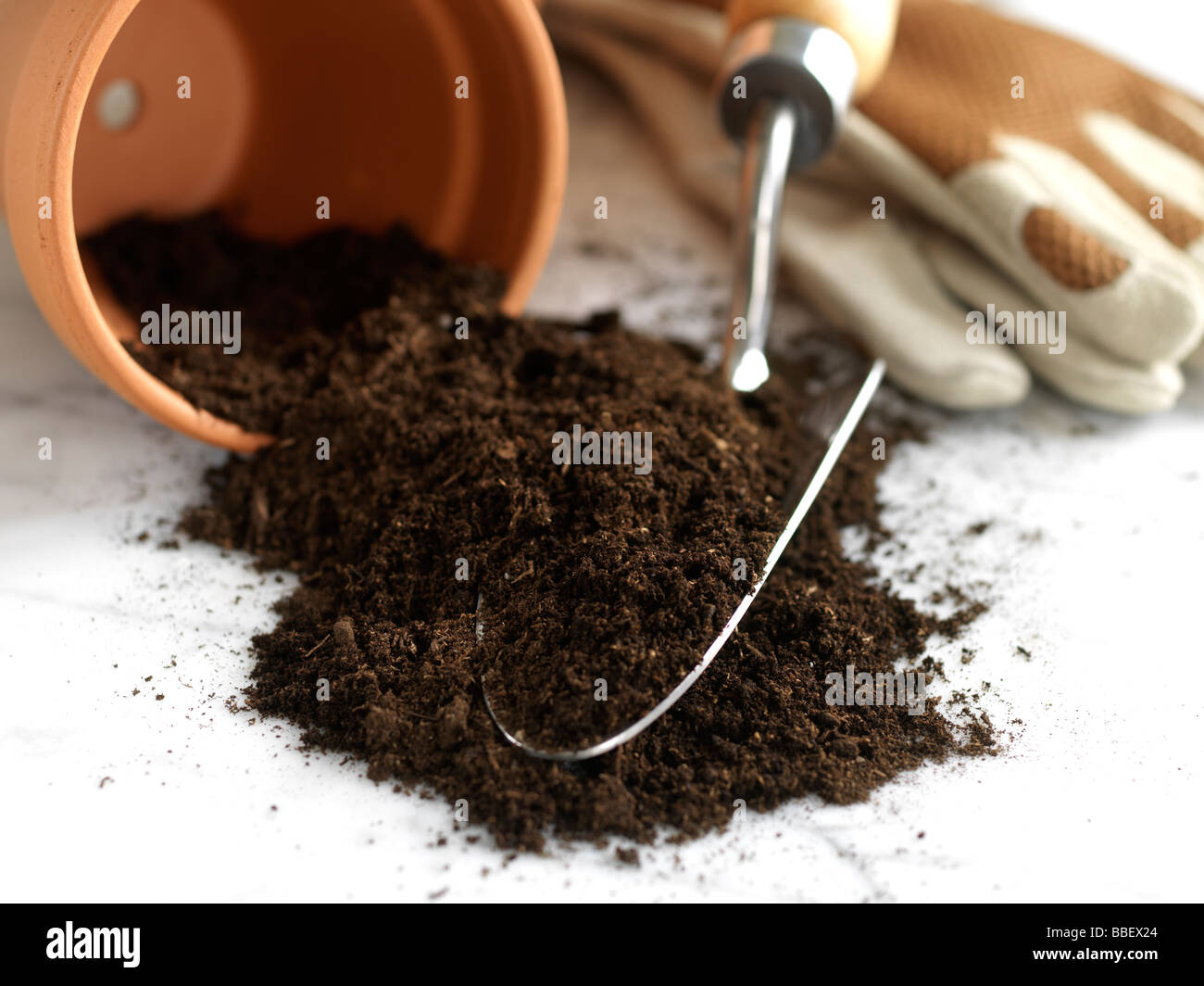 Una maceta de barro con derrames de tierra, una pequeña pala de mano y guantes de jardinería sobre un fondo blanco. Foto de stock