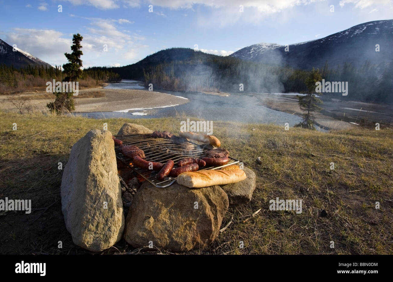 Embutidos, carnes y baguette, sobre un fuego de campamento, barbacoa, cocina, camping, Río Takhini detrás, Territorio de Yukon, Canadá, Norte Foto de stock
