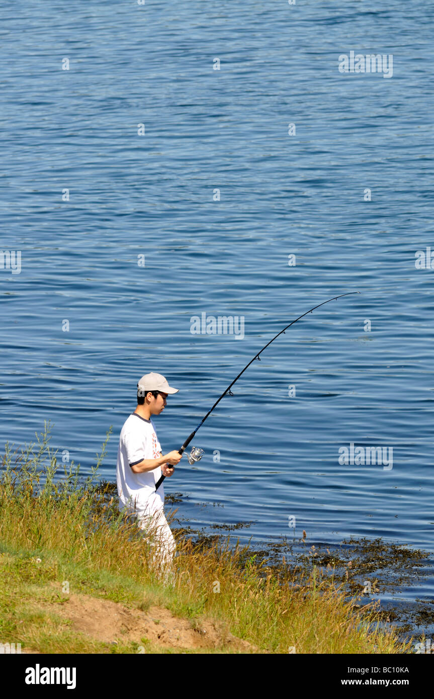 Un adolescente de pesca en el Waters Edge con caña de pescar sombrero y camisa blanca en Canal de Cape Cod en Bourne, Massachusetts Foto de stock