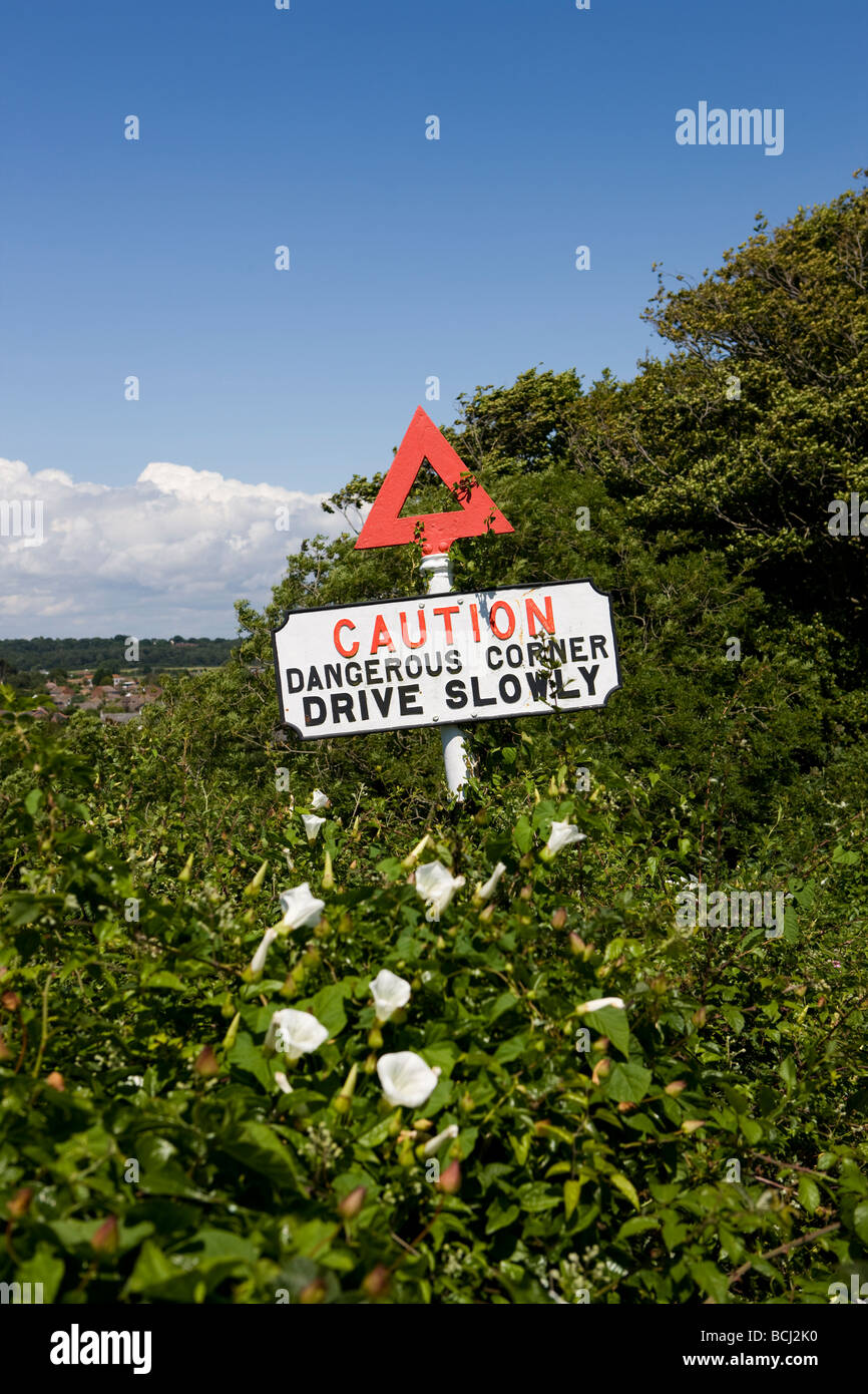 Señal de advertencia en la carretera antigua precaución esquina peligroso conducir lentamente con el triángulo rojo y en los arbustos Foto de stock