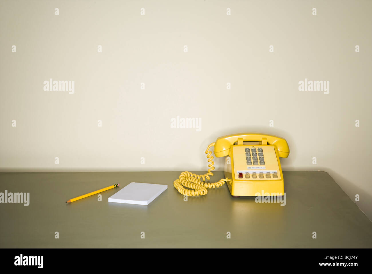 Teléfono de tono amarillo sentado en la mesa metálica delante de la pared blanca. Un lápiz y un bloc de papel sentado junto al teléfono de escritorio Foto de stock