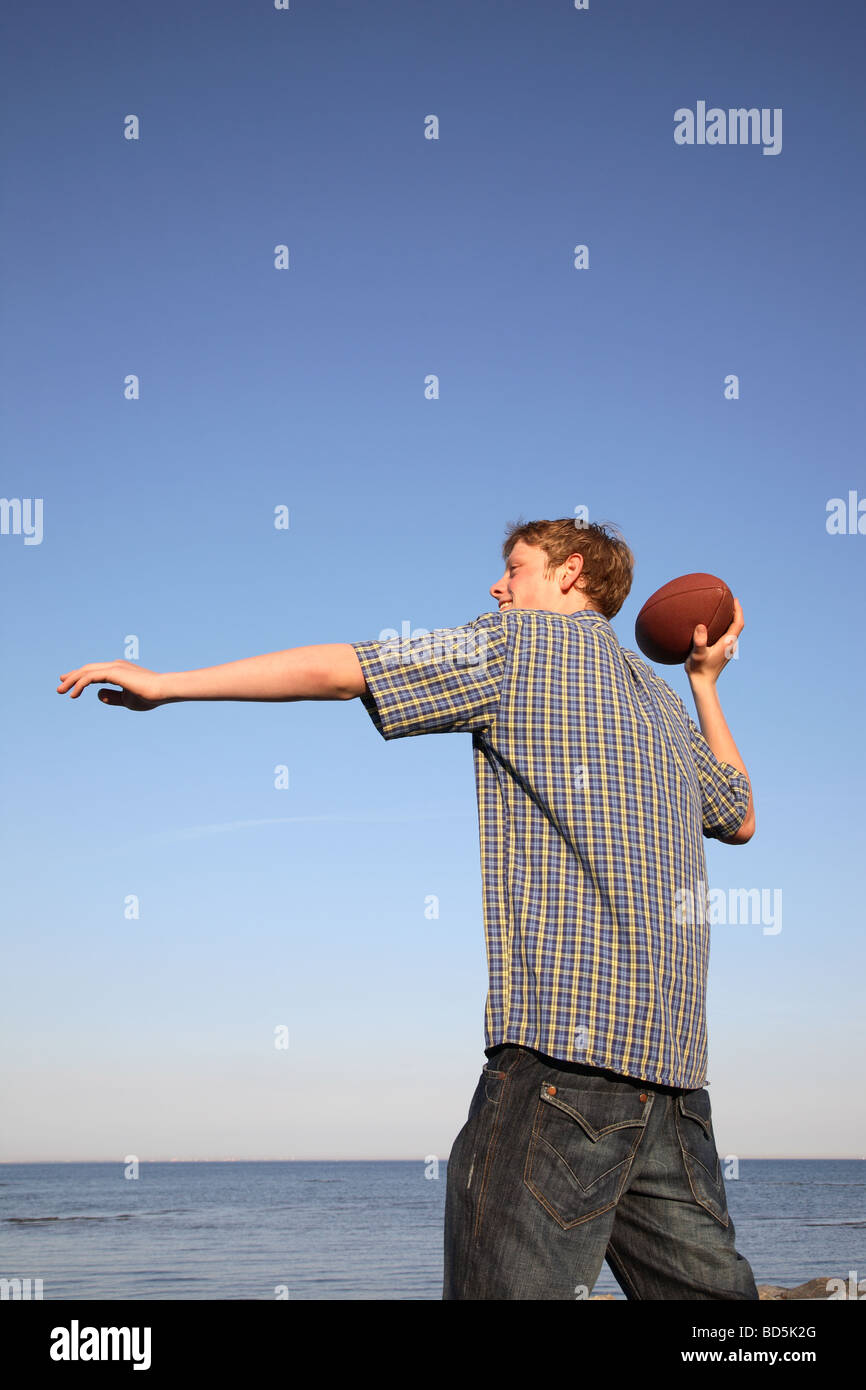 Adolescente lanzando una pelota de fútbol americano en la playa al atardecer contra un cielo azul. Foto de stock