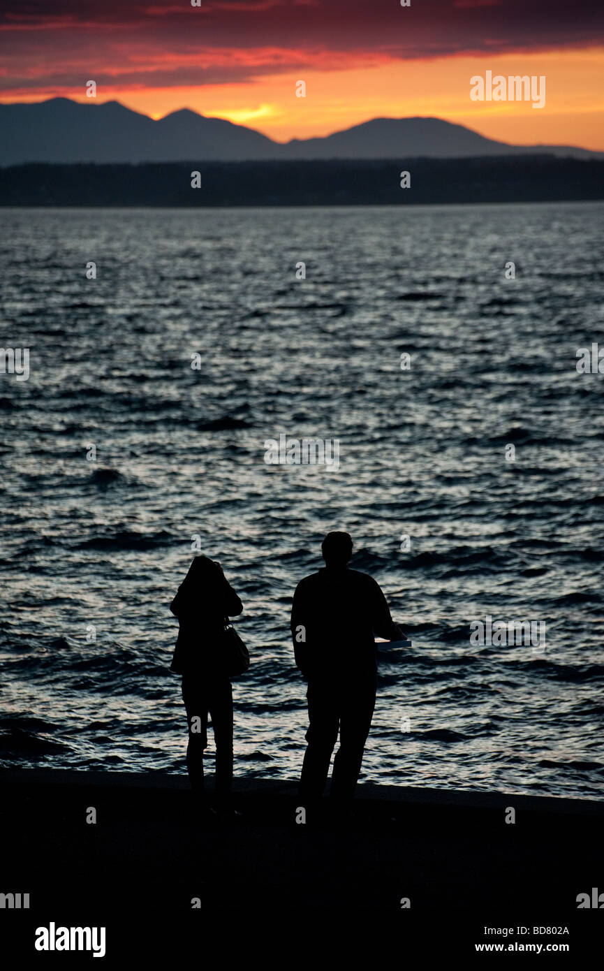 Una joven pareja fotografiar la puesta de sol mientras pasea en Alki Beach. Bainbridge y la Península Olímpica son en el fondo. Foto de stock