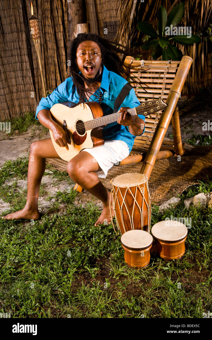 Joven hombre jamaiquino con dreadlocks tocando la guitarra y bongos en isla tropical Foto de stock