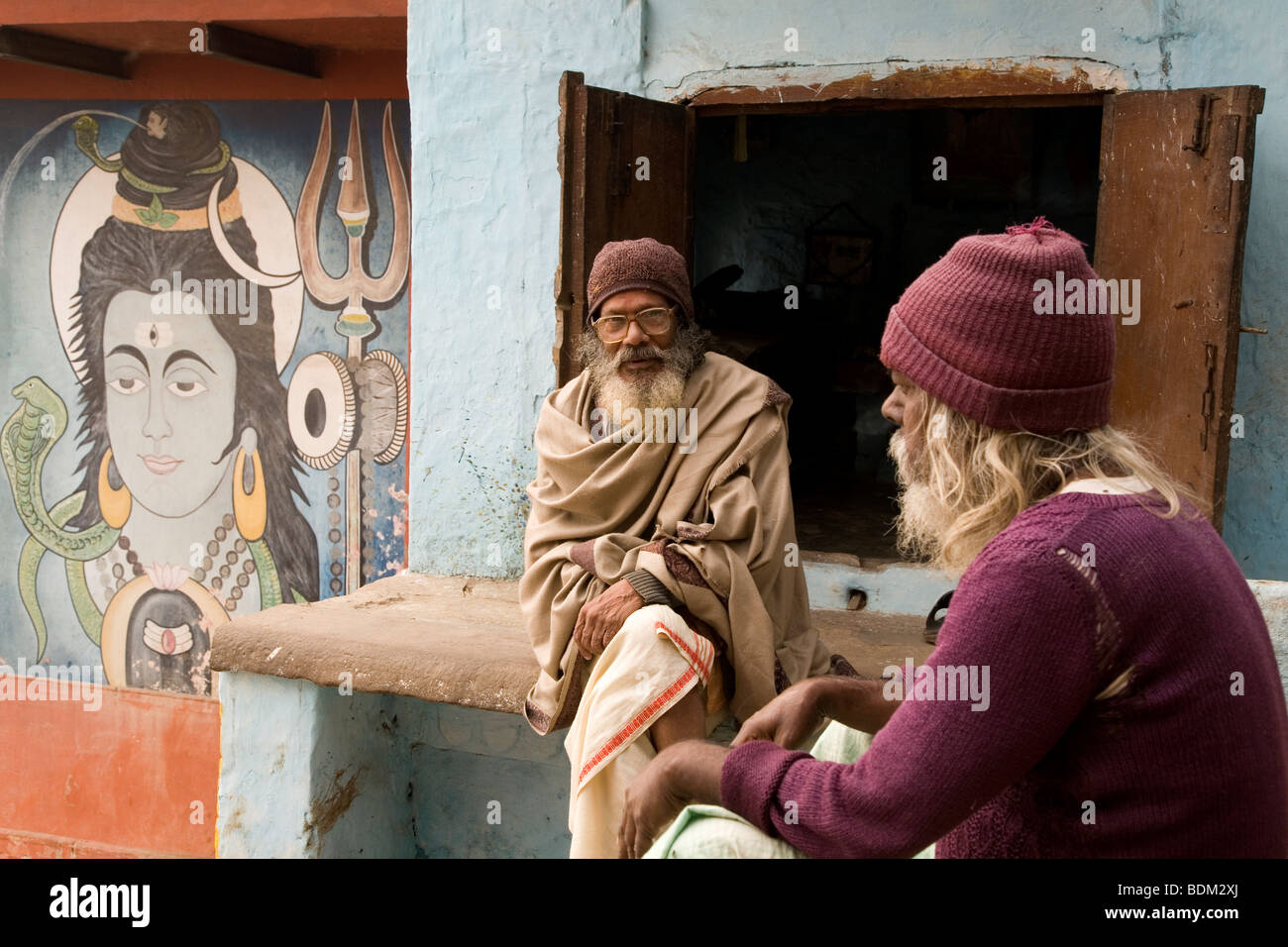 Hombres indios barbudo sentarse y hablar junto a un santuario en Varanasi, India. Una pintura del dios hindú Shiva puede ser visto (izquierda). Foto de stock