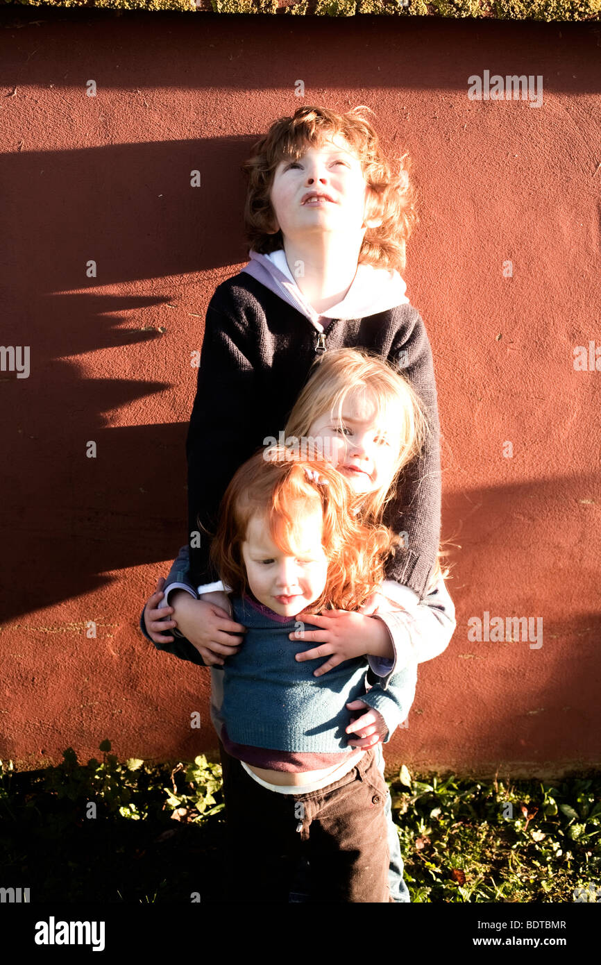 El hermano y las dos hermanas menores de pie contra una pared en la luz del sol Foto de stock
