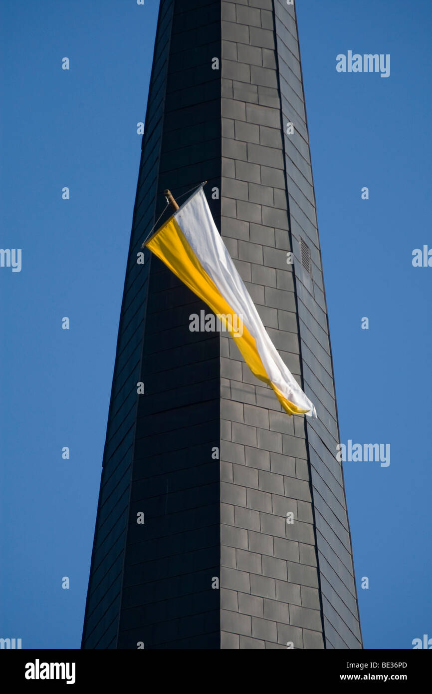 Ondeando la bandera blanca y amarilla desde una ventana de la torre de la iglesia contra un cielo azul acero Foto de stock