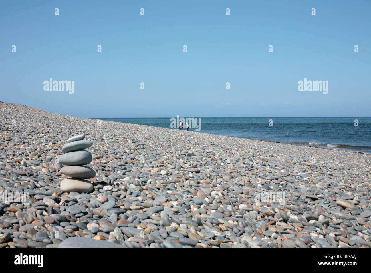 Pila de cinco piedras en el primer plano de una enorme playa de guijarros con tres figuras distantes Foto de stock
