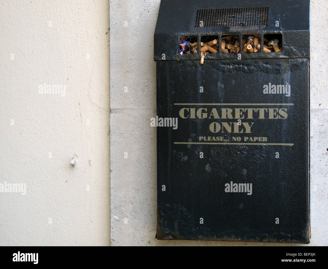 Black metal bin cigarrillos fijada a una pared exterior y un bar lleno con colillas de cigarrillo Foto de stock