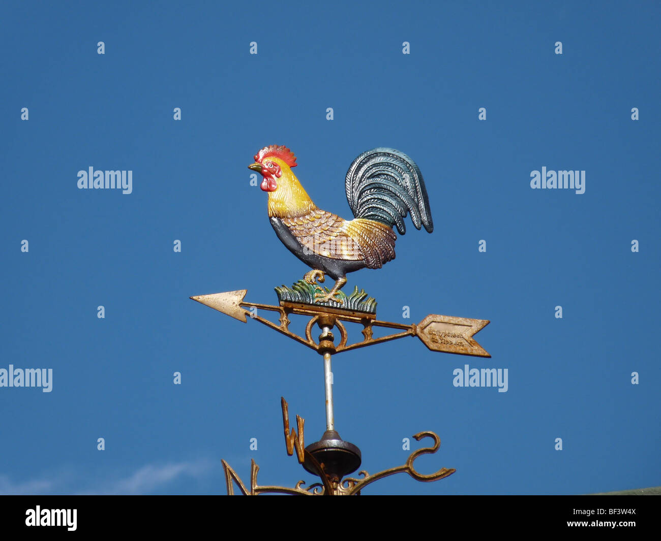 Un metal decorativo de veleta en forma de gallo de colores brillantes, con una flecha apuntando contra un cielo azul brillante Foto de stock