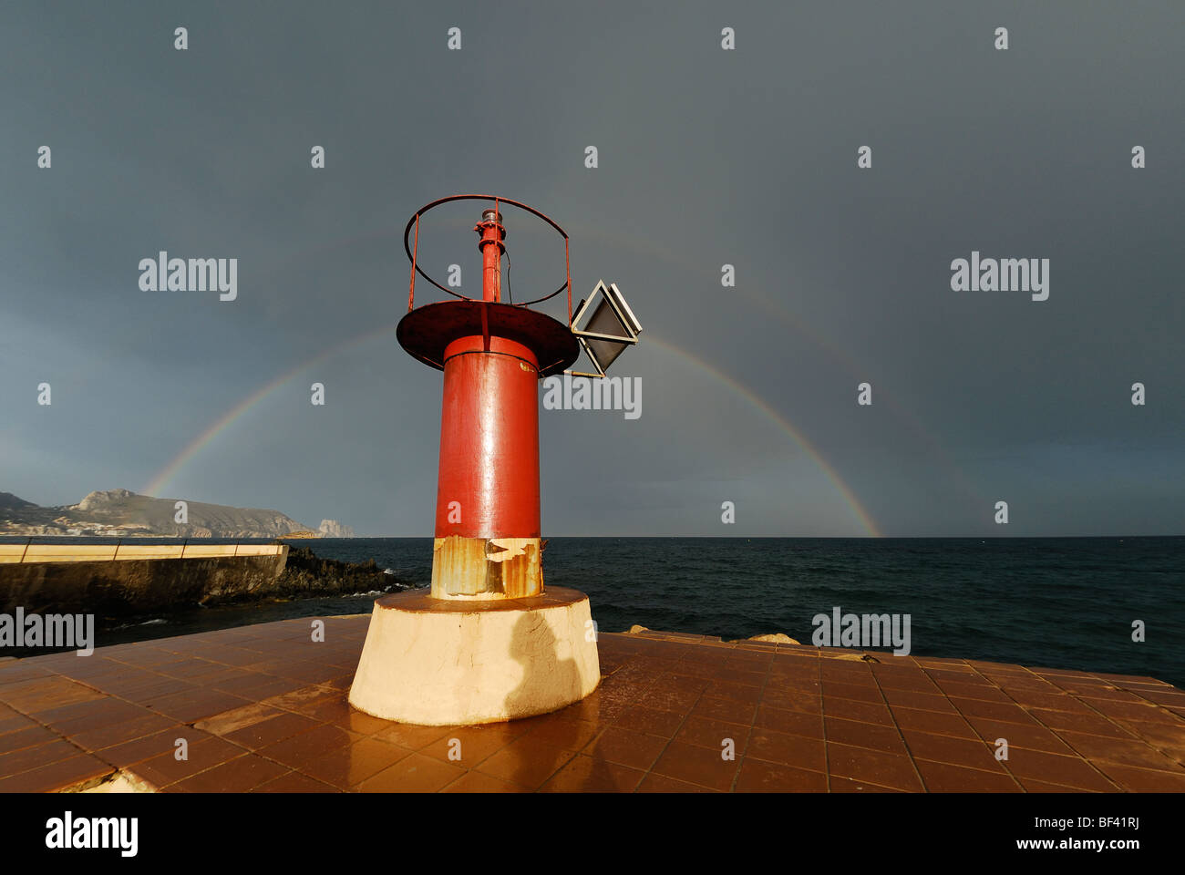 Faro rojo en el muelle de puerto pequeño en frente de nubes de tormenta y Rainbow en Cap Negret, Altea, Costa Blanca, España Foto de stock