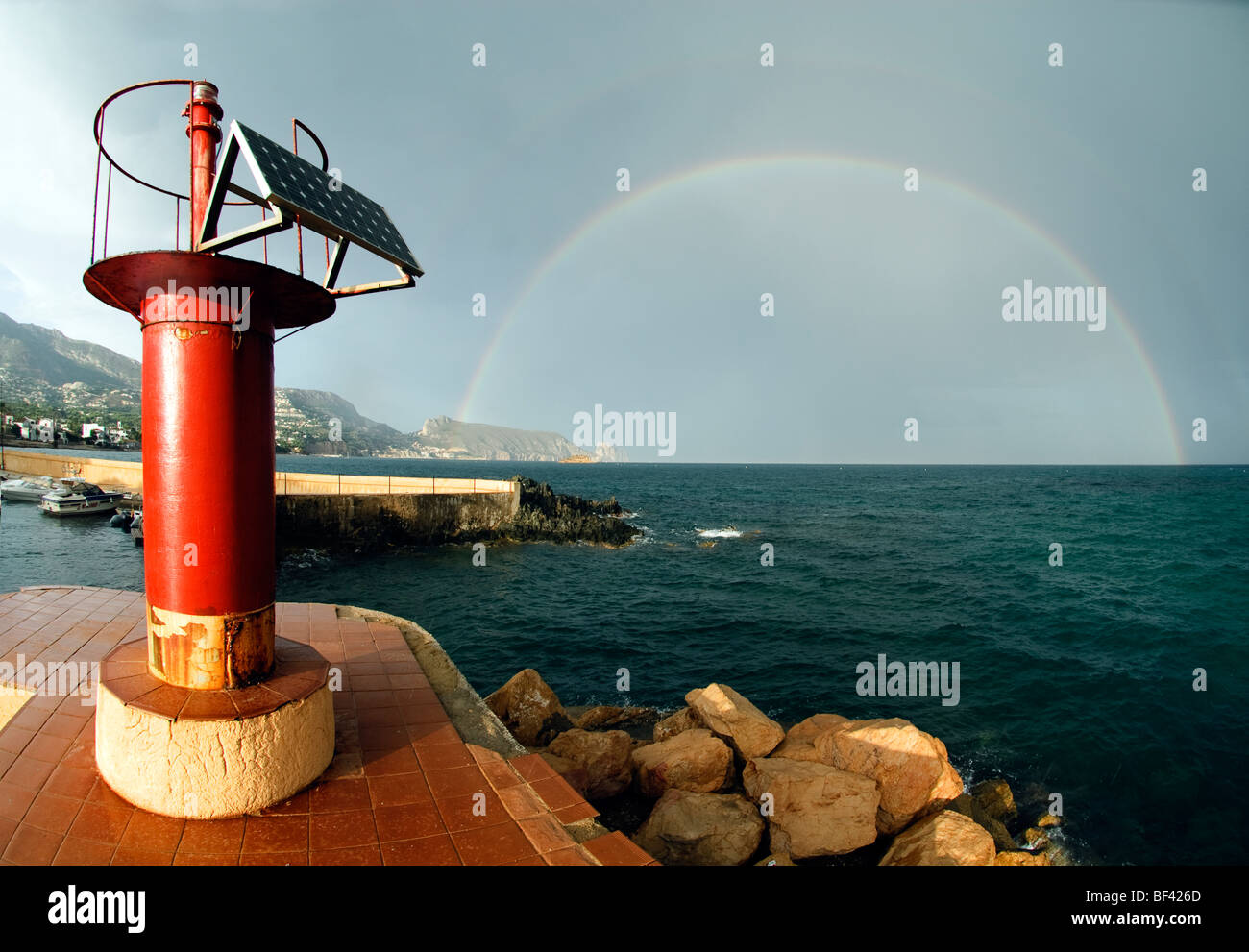 Faro rojo en el muelle de puerto pequeño en frente de nubes de tormenta y Rainbow en Cap Negret, Altea, Costa Blanca, España Foto de stock
