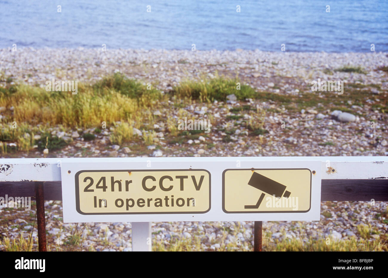 Playa de grava ásperos con hierbas y mar con oxidada puerta e icónica imagen de cámara CCTV de 24hr de advertencia y en funcionamiento Foto de stock