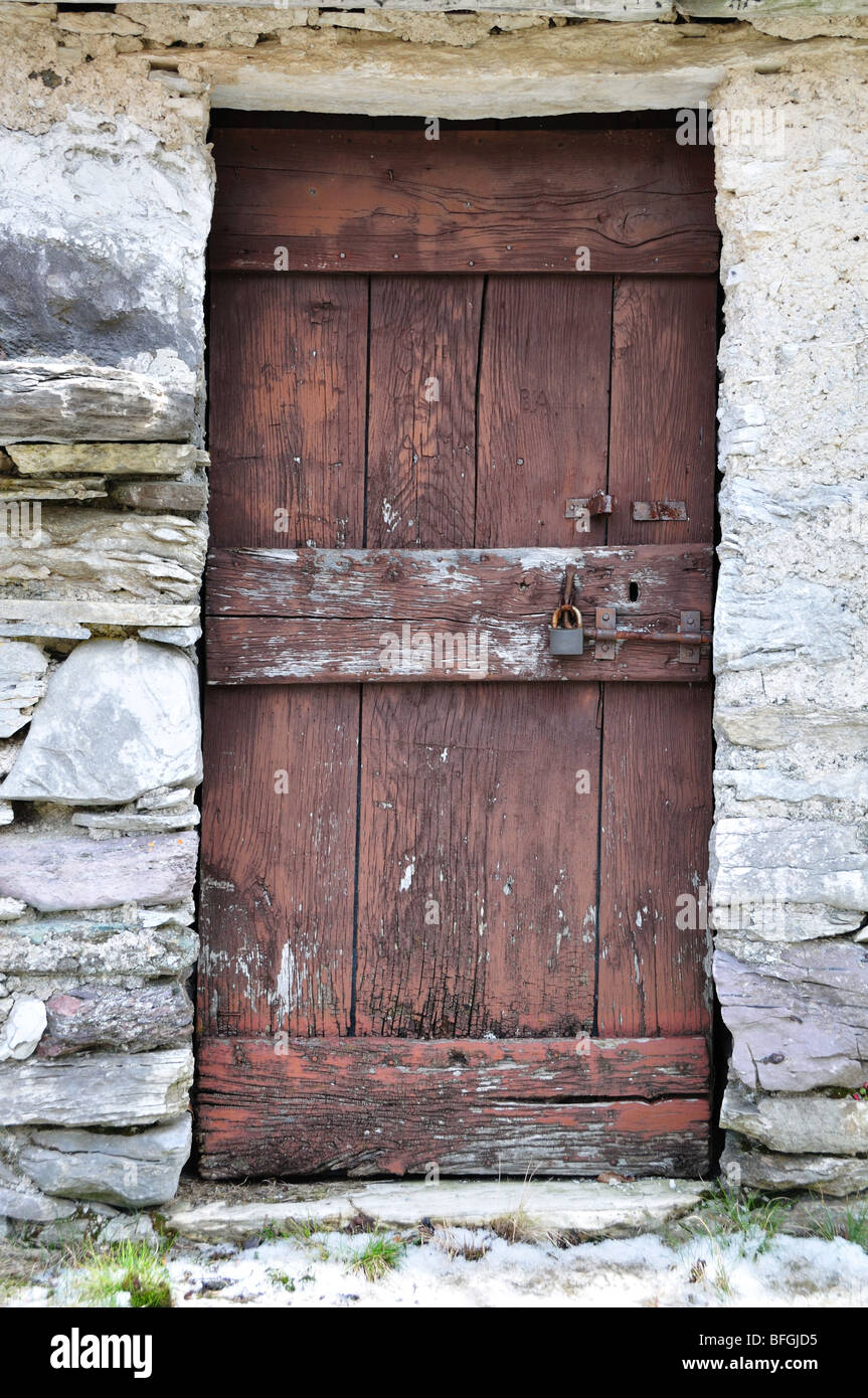 Puerta de madera vieja en un muro de piedra de un antiguo edificio abandonado de un pueblo de montaña en los Alpes Italianos Foto de stock