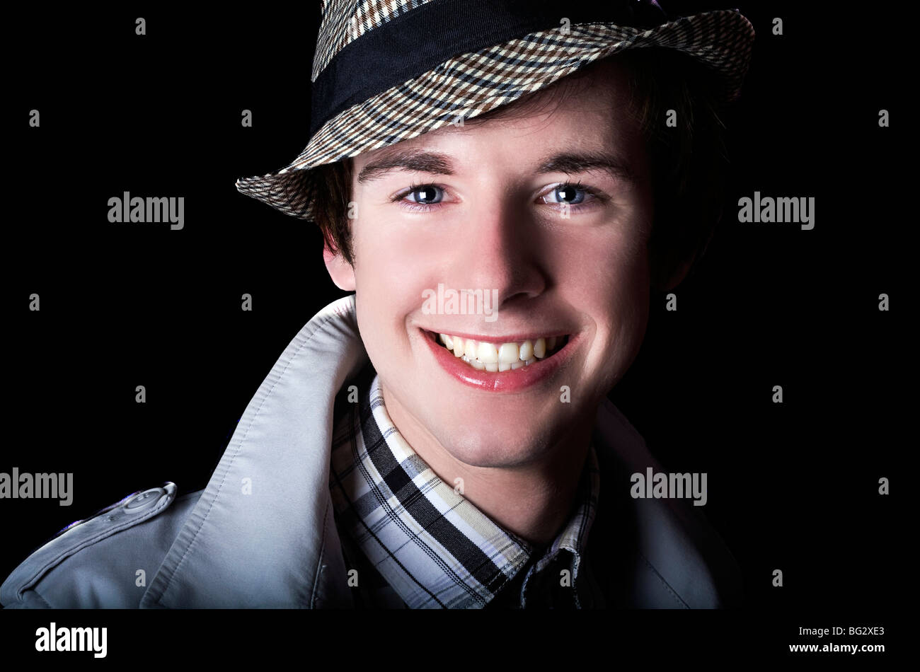 Un hombre joven de moda en un impermeable y trilby contra un escenario dramático oscuro Foto de stock
