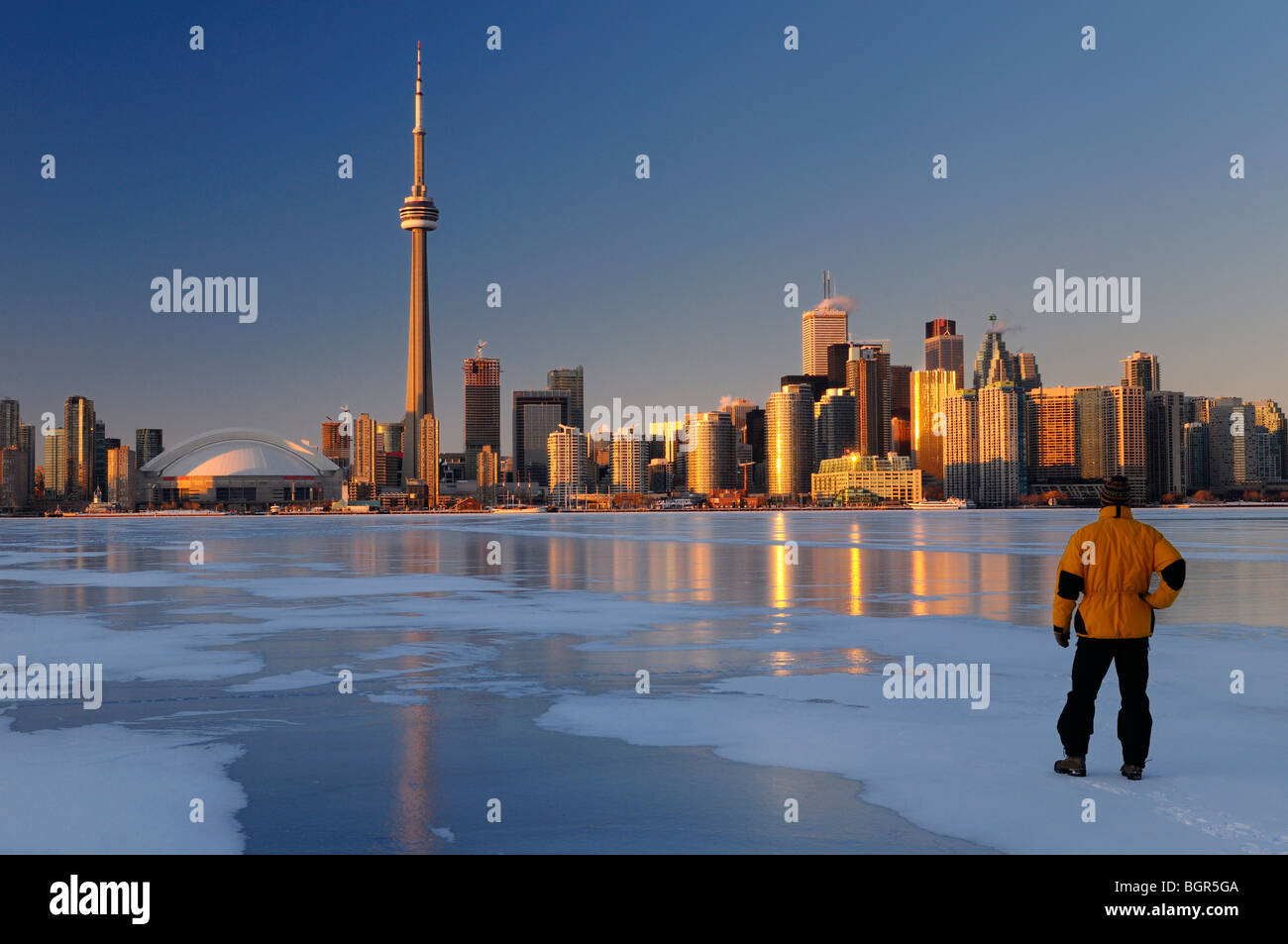 Hombre de pie sobre el hielo del lago Ontario congelado mirando al horizonte de la ciudad de Toronto con brillo dorado al atardecer Foto de stock