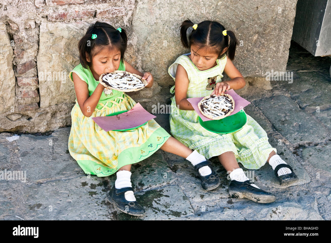 Dos niñas mexicanas pequeñas en bonitos vestidos - cuadros & rayados Merceditas sentarse en la acera y disfrutar del almuerzo trata de tacos idénticos Foto de stock