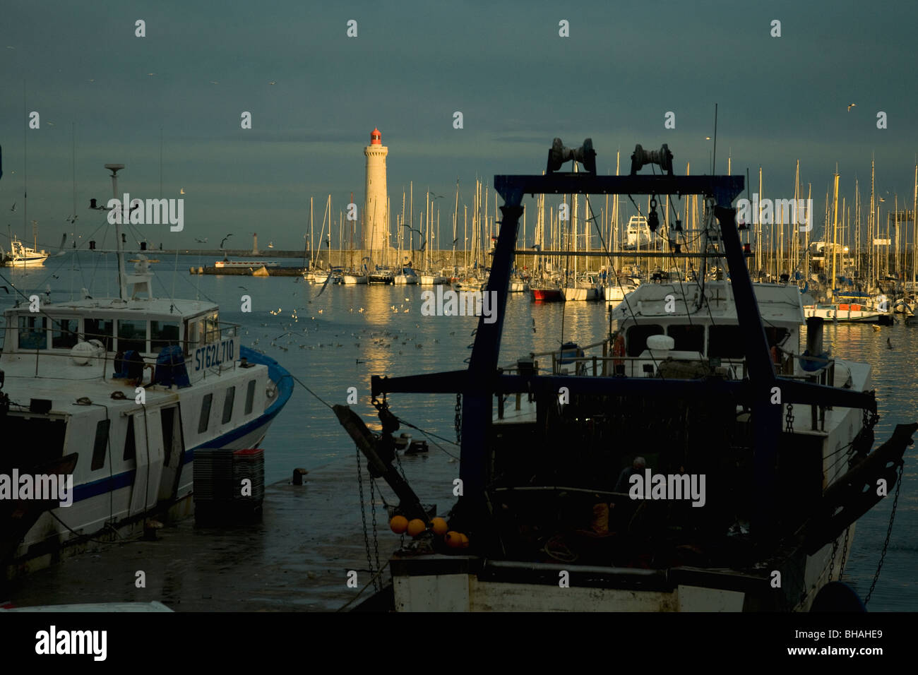 En Sète, Francia, el mayor puerto pesquero del Mediterráneo, los arrastreros están amarrados tranquilamente como el sol vespertino ilumina el faro Foto de stock
