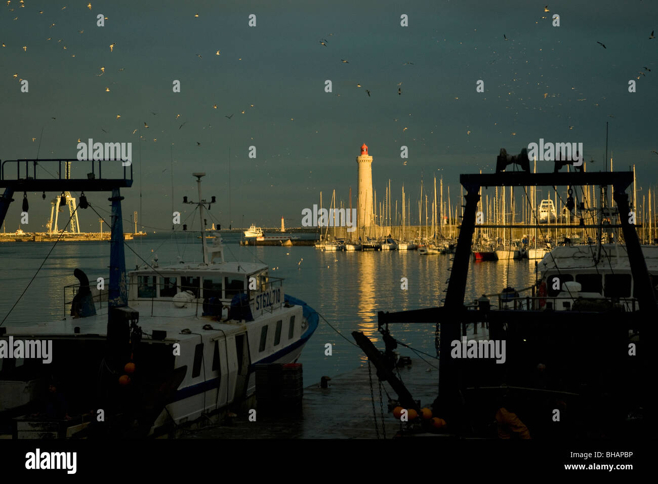En Sète, Francia, el mayor puerto pesquero del Mediterráneo, los arrastreros están amarrados tranquilamente como el sol vespertino ilumina el faro Foto de stock
