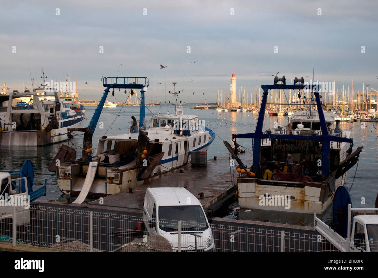 En Sète, Francia, el mayor puerto pesquero del Mediterráneo, los arrastreros están amarrados tranquilamente como el sol vespertino ilumina el faro Foto de stock