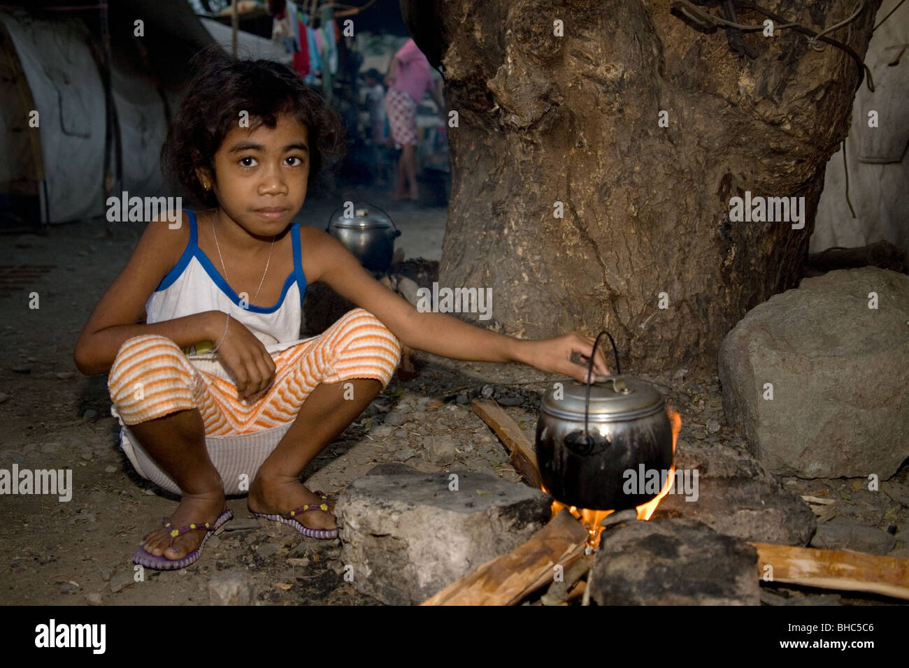 Mujer joven con olla a fuego abierto prepara una comida en el campamento para desplazados de IDP abarrotado de familias sin hogar en Dili, Timor Oriental Foto de stock