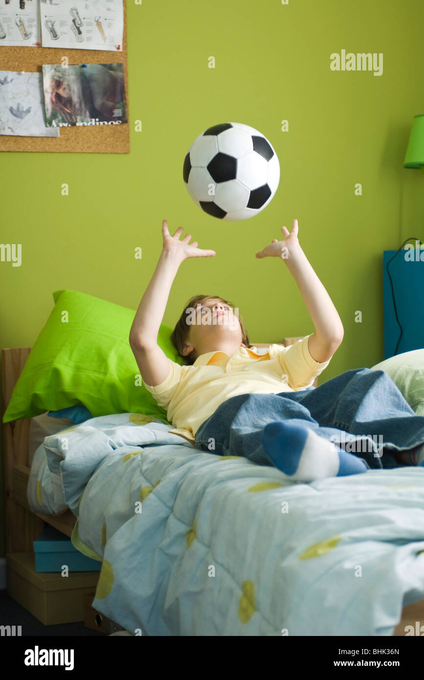 Niño acostado en la cama de brazos cruzados arrojando la bola en el aire Foto de stock
