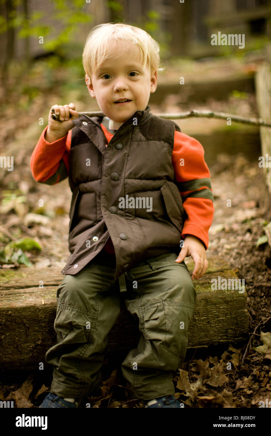 Muchacho sentado en el bosque sosteniendo un palo. Foto de stock