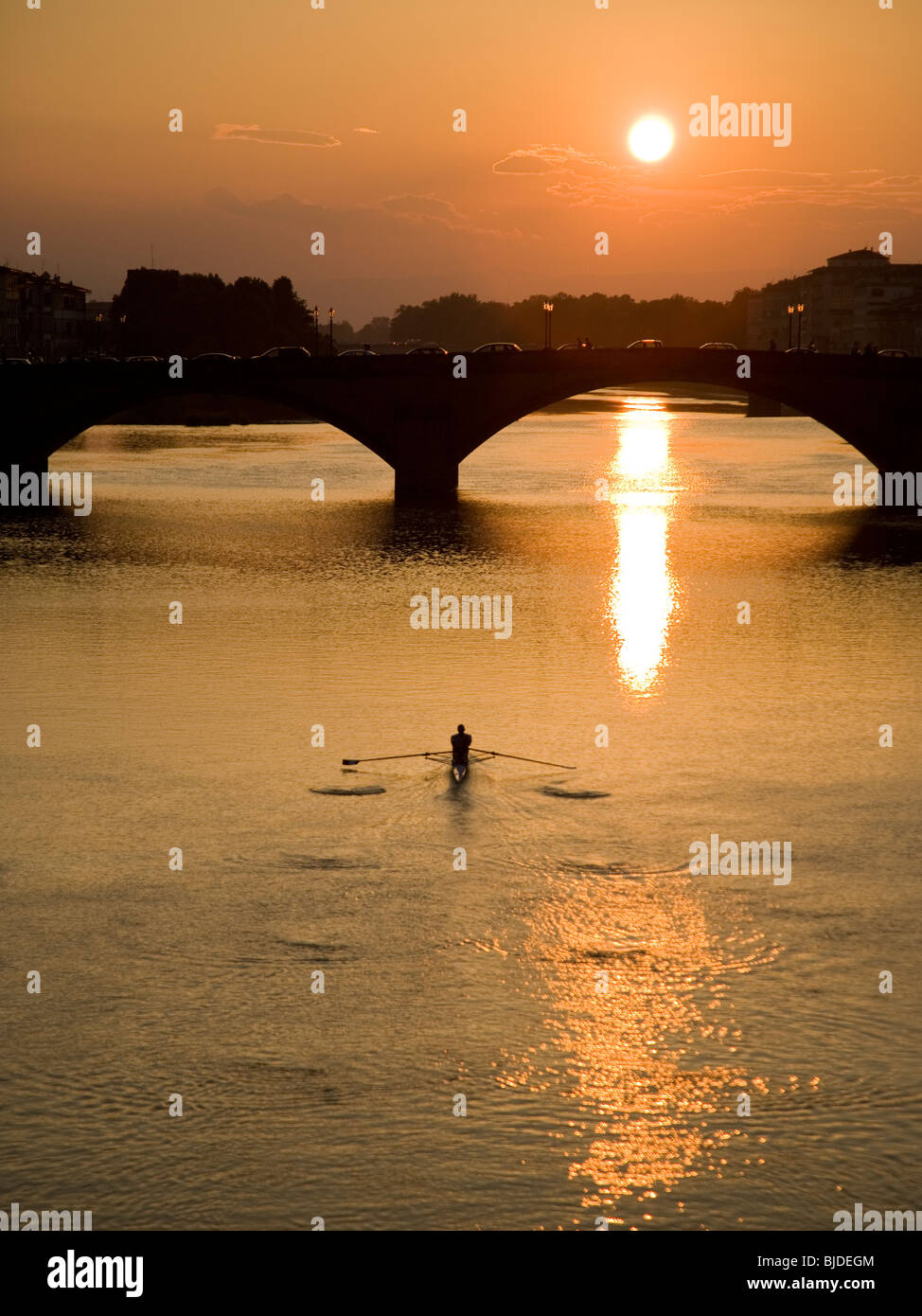 Hombre remando en un cuerpo de agua con un puente y una puesta de sol en el fondo. Foto de stock