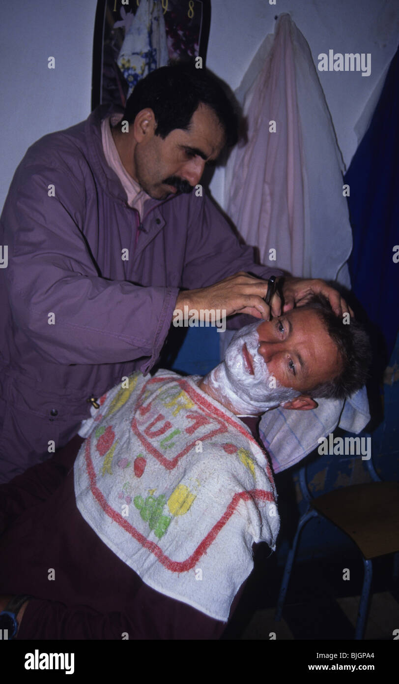 Hombre sentado en una silla, habiendo un afeitado húmedo tradicional con una hoja de afeitar por un barbero, Marruecos. Foto de stock