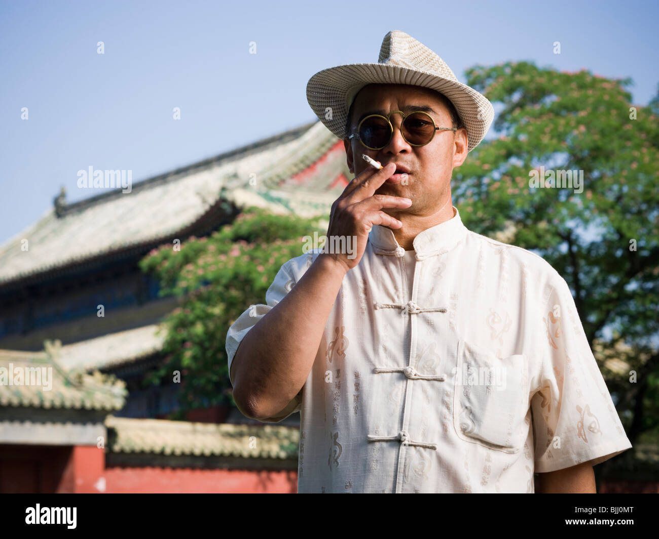 Hombre con gafas de sol y sombrero de paja de fumar cigarrillo afuera con pagoda en segundo plano. Foto de stock