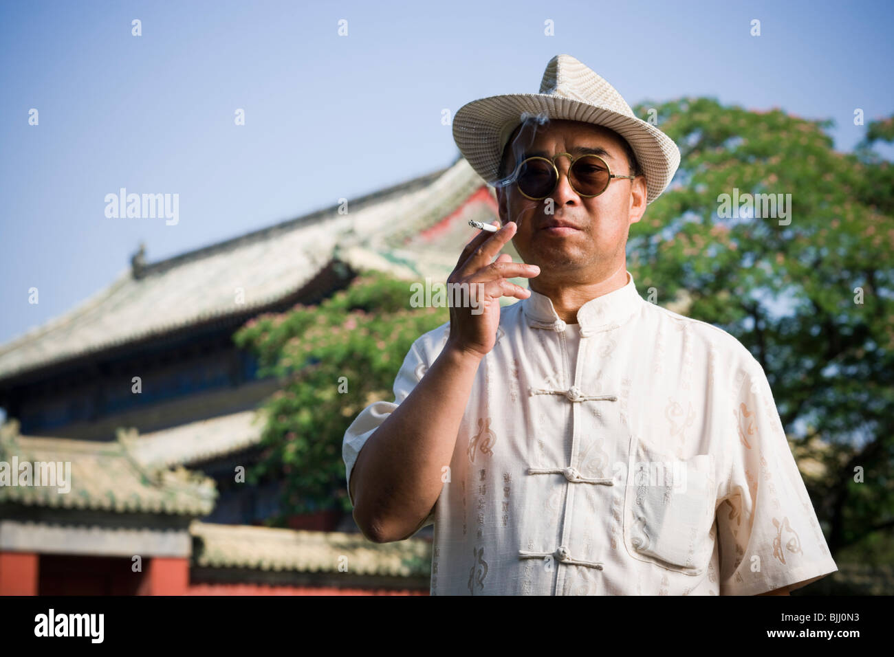Hombre con gafas de sol y sombrero de paja de fumar cigarrillo afuera con pagoda en segundo plano. Foto de stock