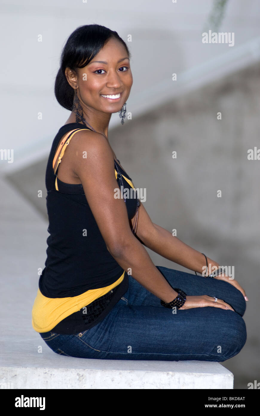 Un joven americano africano adulto hembra está sentado en un muro de hormigón sonriendo Foto de stock
