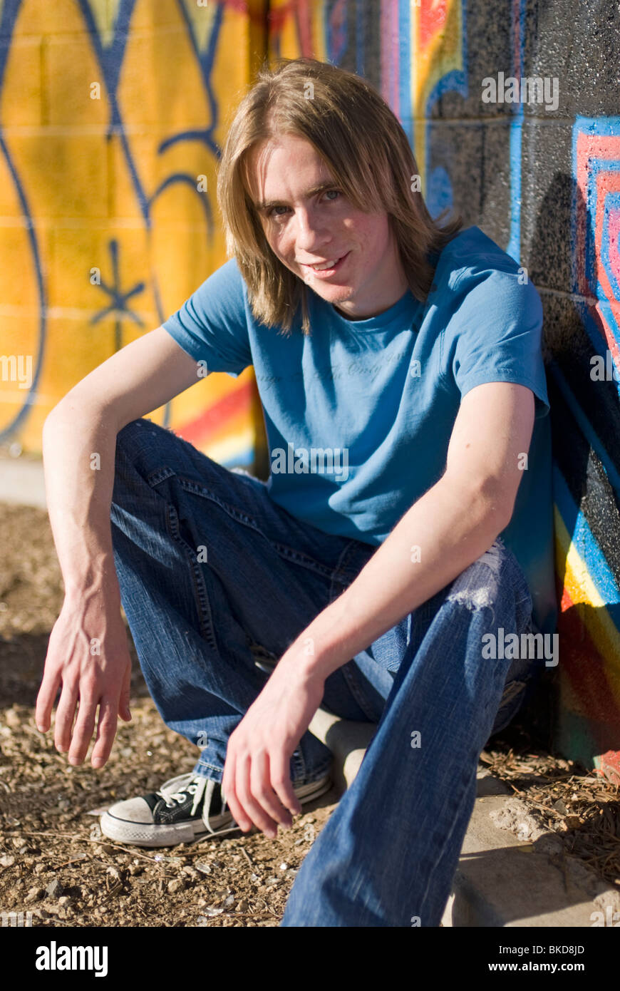 18-20 años de edad, hombre en zapatillas, pantalones vaqueros y una camiseta de cuclillas cerca de una pared cubierta de graffiti - sonriendo (vertical) Foto de stock