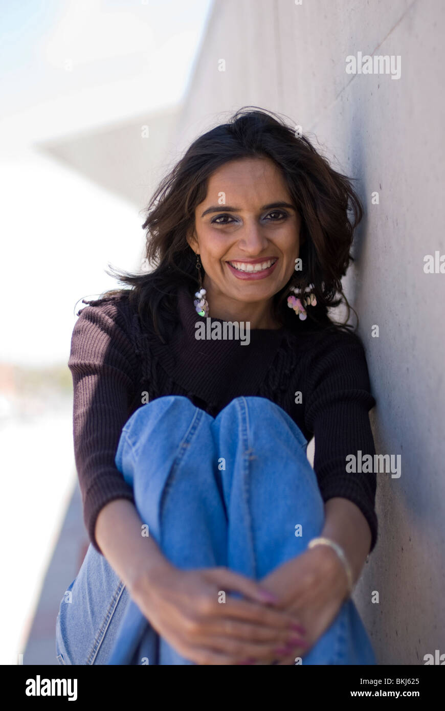 Una joven y bella mujer india (Oriental) se sienta y se inclina sobre una pared de color claro - ella está sonriendo. Foto de stock