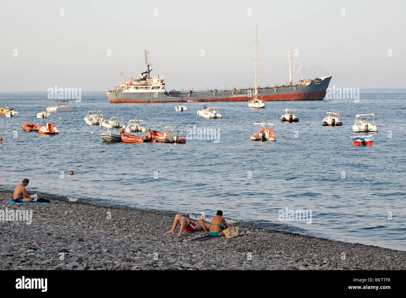 Turistas sentados en una playa rocosa con barcos en el fondo en Santa Marina, en la isla de Salina, Islas Eolias, Sicilia, Italia. Foto de stock