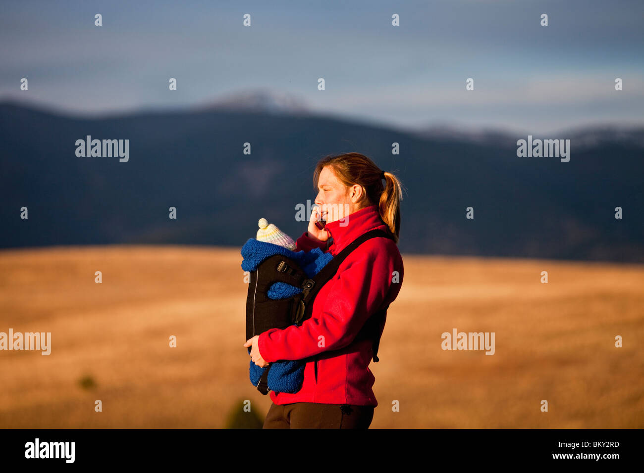 Madre en el teléfono celular y el recién nacido en el soporte delantero mientras senderismo por las colinas cubiertas de hierba, Obras Hidráulicas, Missoula, Montana. Foto de stock