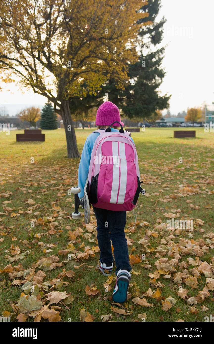 Una chica joven llevando su monopatín, camina a casa desde la escuela en un día de otoño. Foto de stock