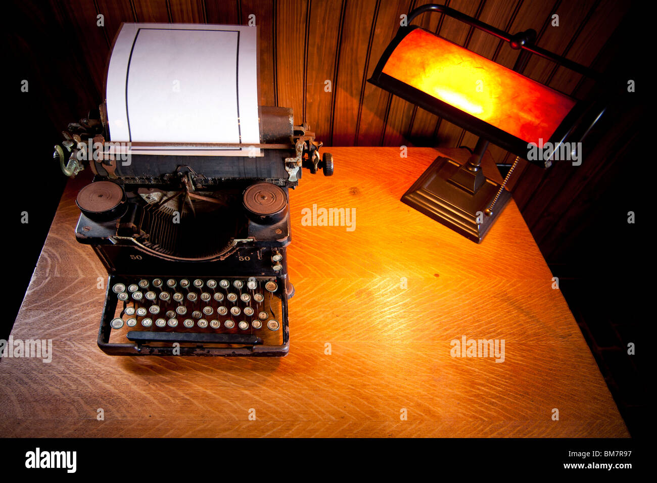 Escritorio de madera iluminada por una lámpara vieja con luz amarilla y un histórico rusty typewriter plastificado con una hoja de papel en blanco. Foto de stock