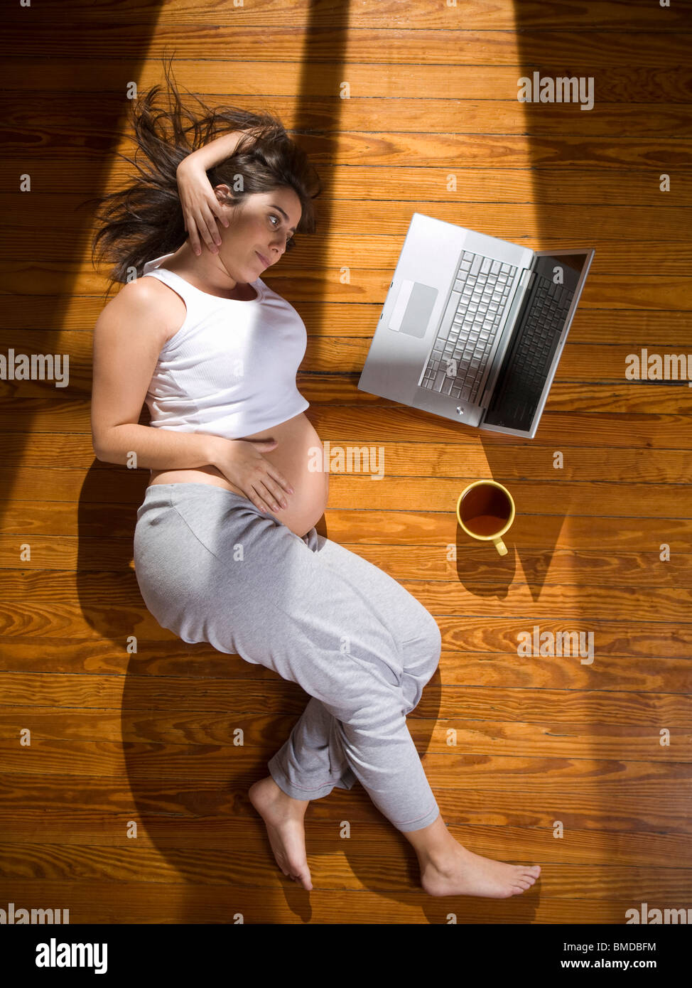 Vista superior de una joven embarazada, tumbado en el suelo con un ordenador portátil y una taza de té. Foto de stock