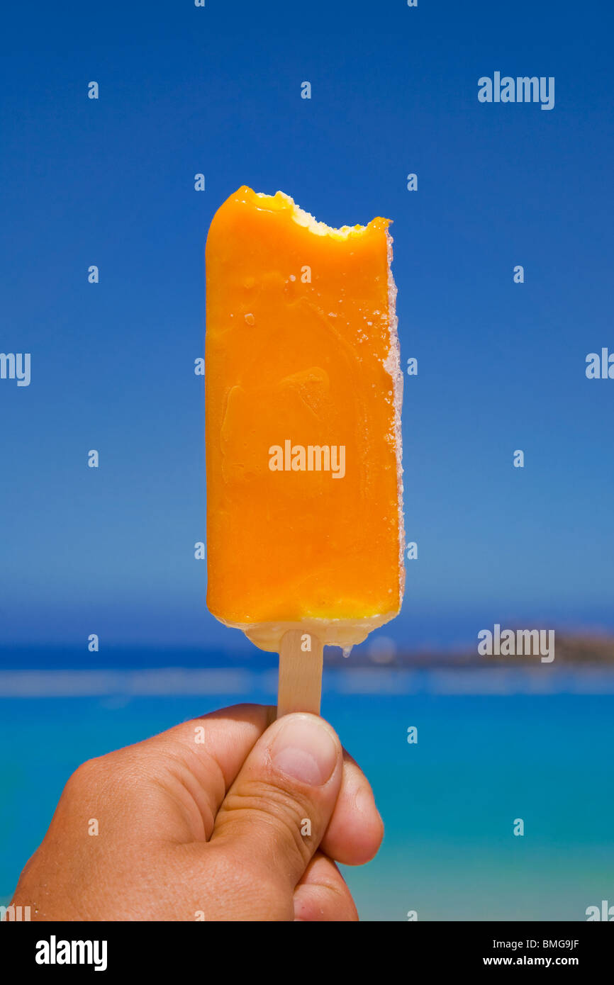 Máquina de paleta de helado en la playa; mano sosteniendo una paleta congelada delante del hermoso cielo azul y verde del agua Foto de stock