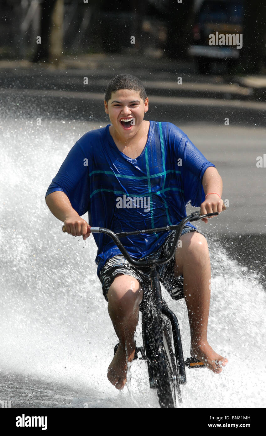 Un adolescente joven monta su bicicleta a través de una corriente de agua de una boca de incendios durante una ola de calor en el CT USA Foto de stock