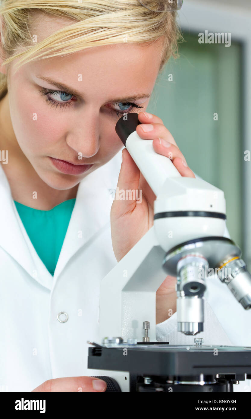 Una hermosa rubia de ojos azules, científico médico femenino o mujer investigador científico utilizando su microscopio en un laboratorio. Foto de stock