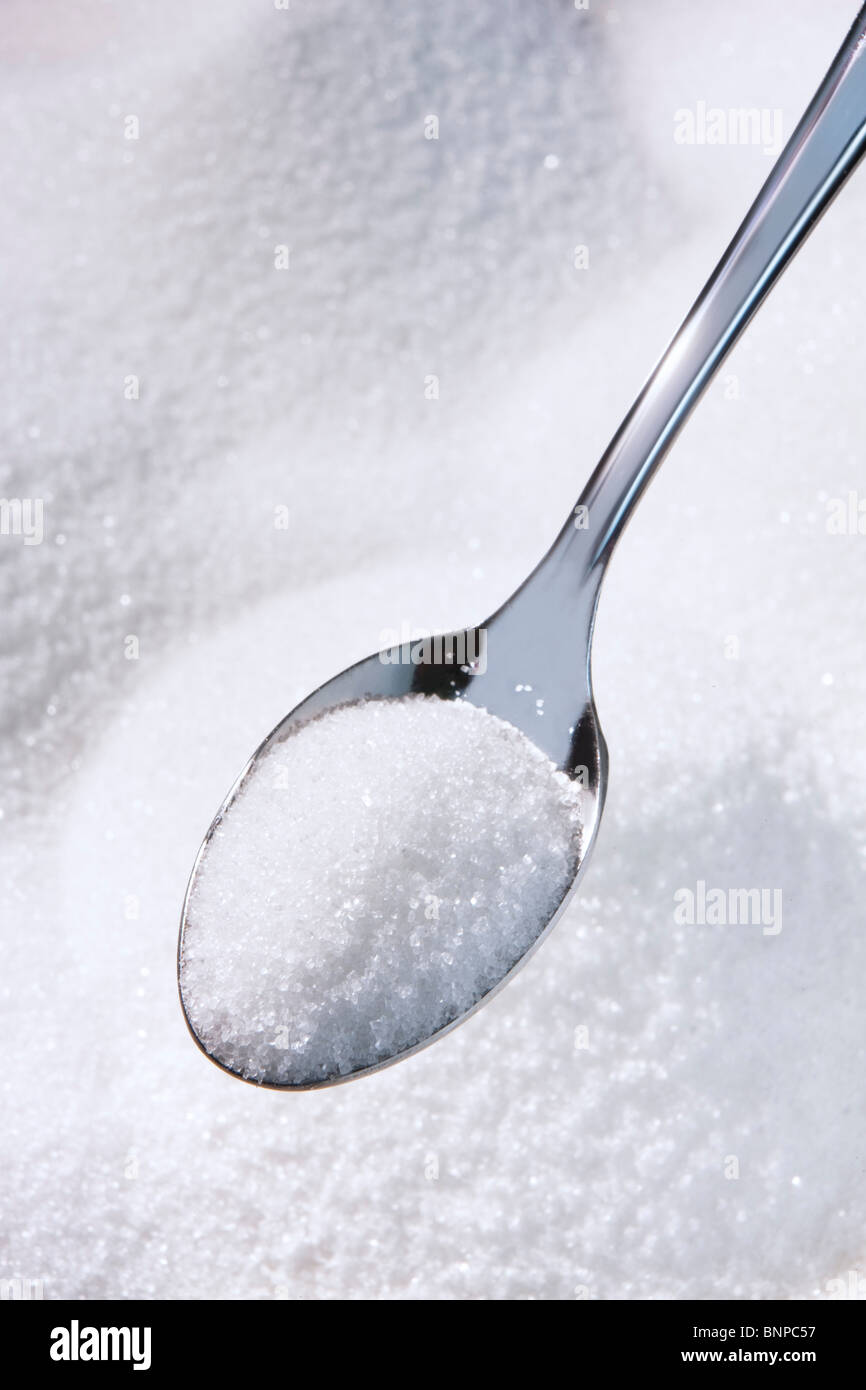 Azúcar blanco granulado en una cucharadita, montones de azúcar en el fondo Foto de stock