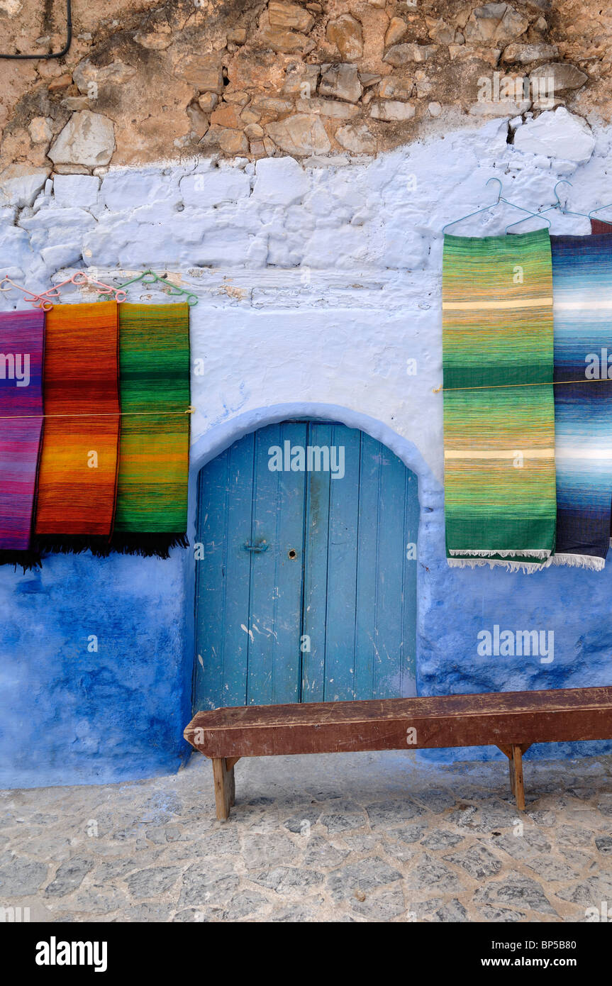 Visualización de coloridas alfombras tejidas, colgantes alfombras o moquetas en venta, Puerta Azul y banco de madera, Chefchaouen, Marruecos Foto de stock