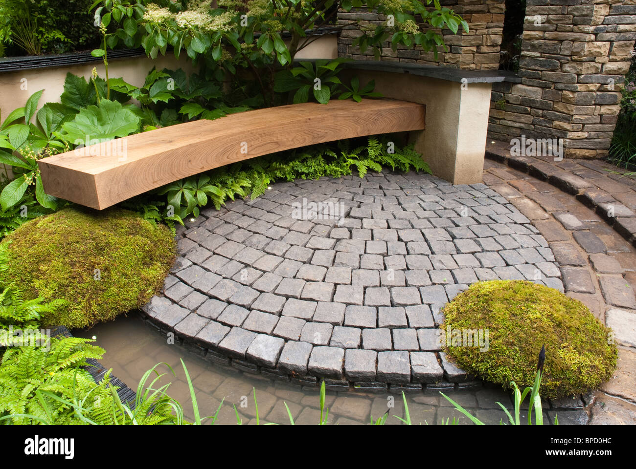 Banco de jardín y patio de piedra circular en curvas, con MOSS, pared, setos, helechos Foto de stock
