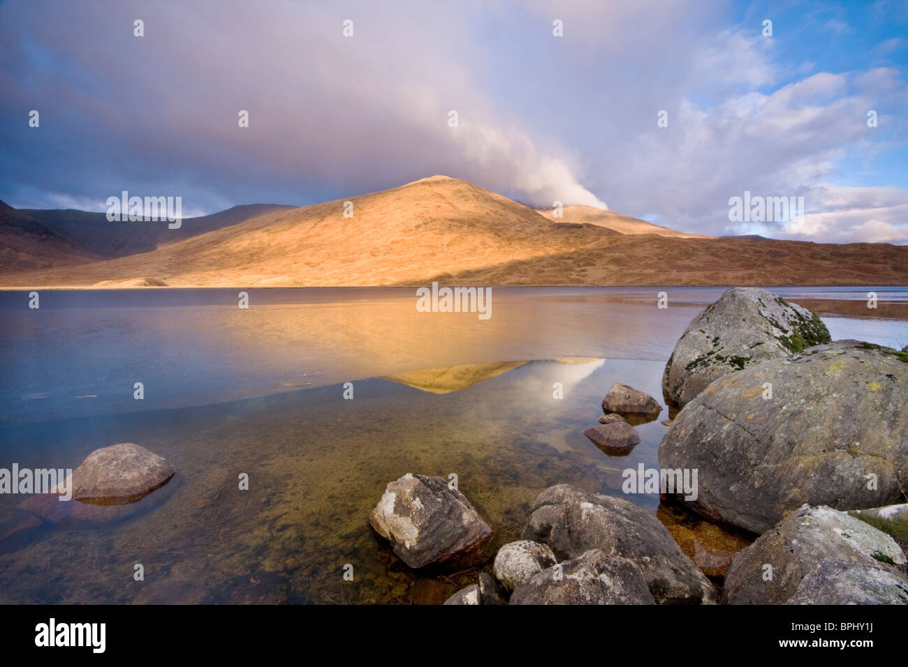 Vista iluminada por el sol de invierno y Araich Meall un Loch Dochard, Argyll and Bute, en Escocia con cantos rodados en primer plano y el hielo sobre el lago Foto de stock